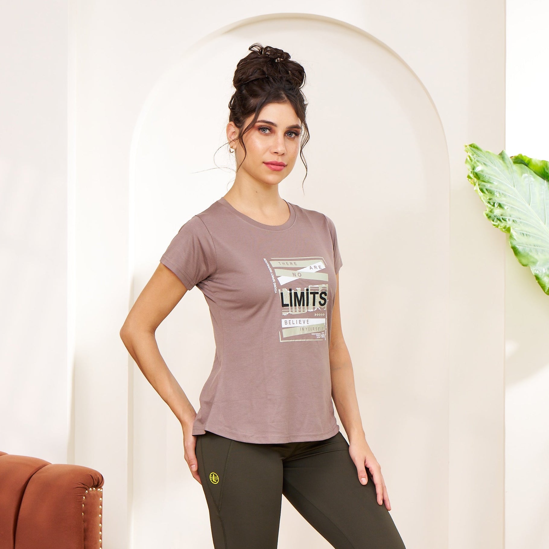 Woman wearing a brown t-shirt and black leggings standing in a room with a plant and red couch.