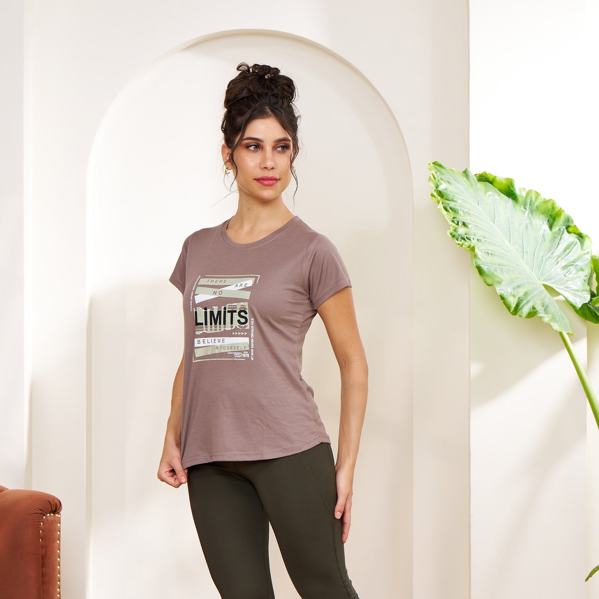 Woman standing in a room with a plant and a couch, wearing a t-shirt with text and leggings.