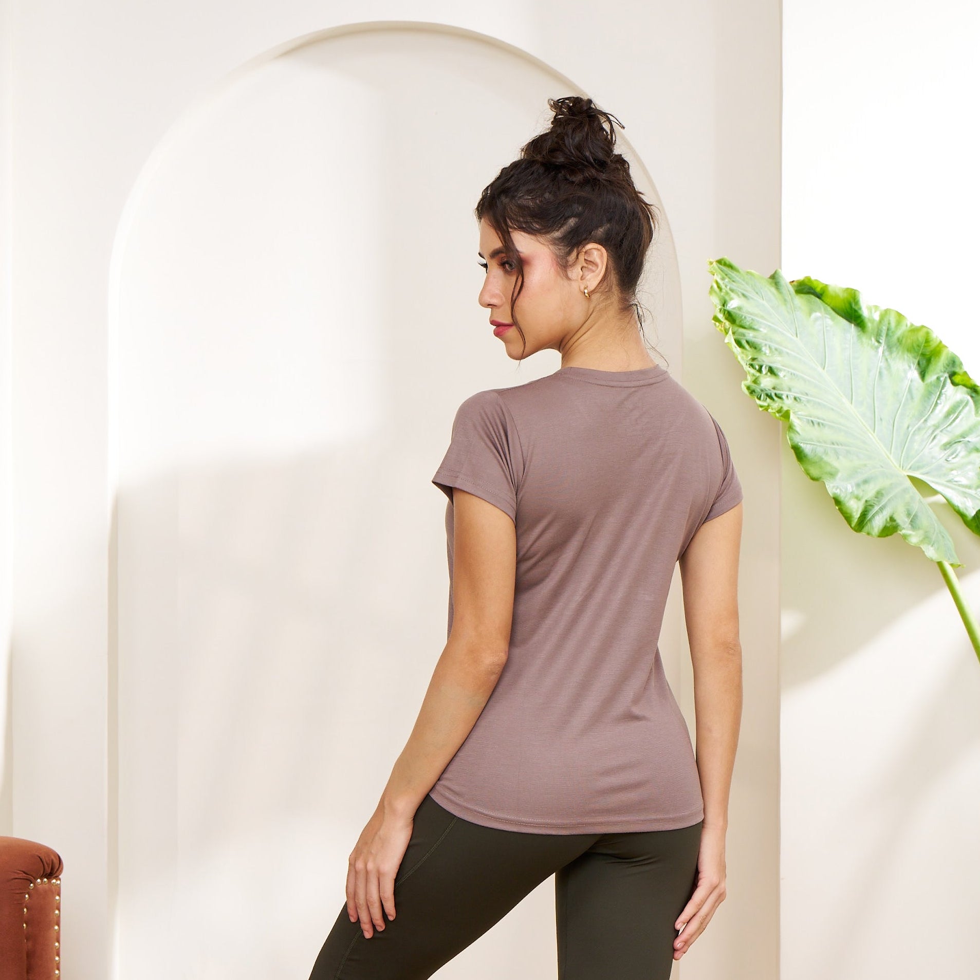 Woman in a brown t-shirt and black leggings standing in a room with a plant and rug.