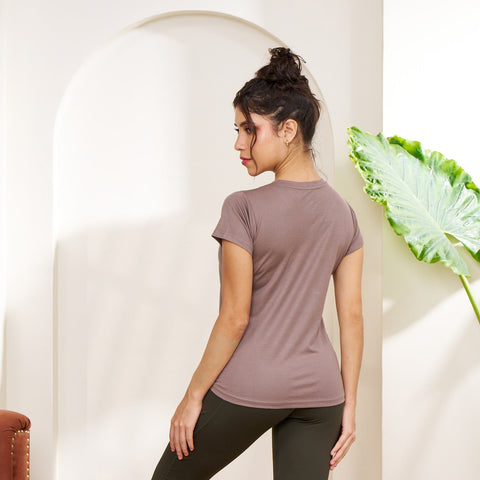 Woman in a brown t-shirt and black leggings standing in a room with a plant and rug.
