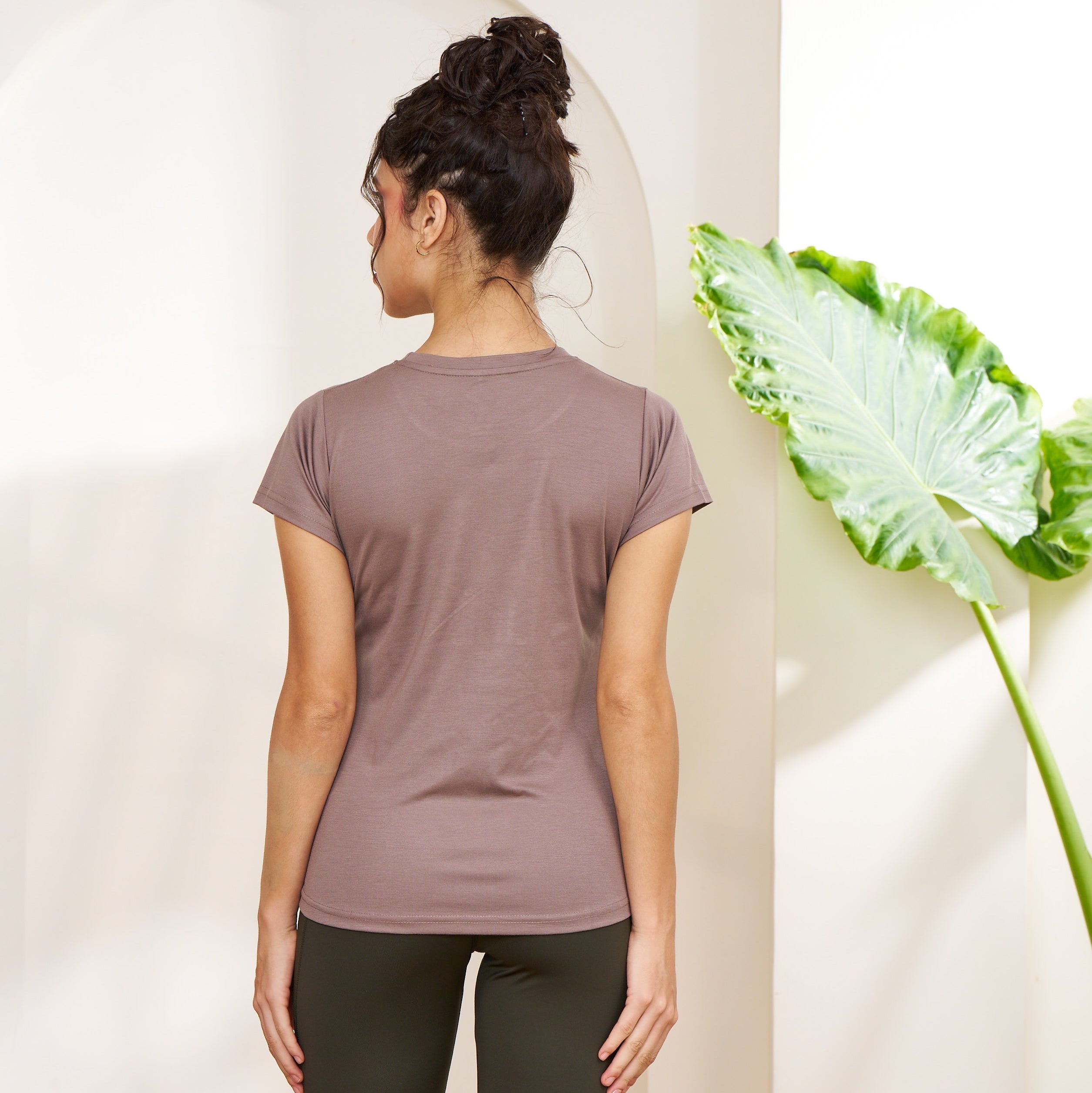 Person wearing a mauve t-shirt and dark pants standing against a white wall with a plant on the right.