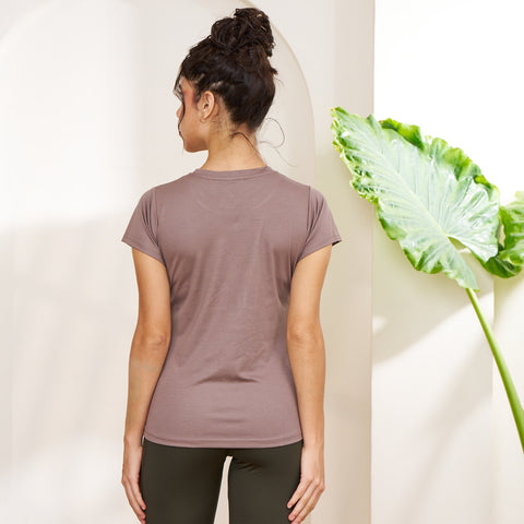 Person wearing a mauve t-shirt and dark pants standing against a white wall with a plant on the right.