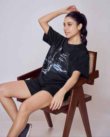 Woman sitting on a wooden chair wearing a black t-shirt and shorts against a plain background. Co-ord Set for Women