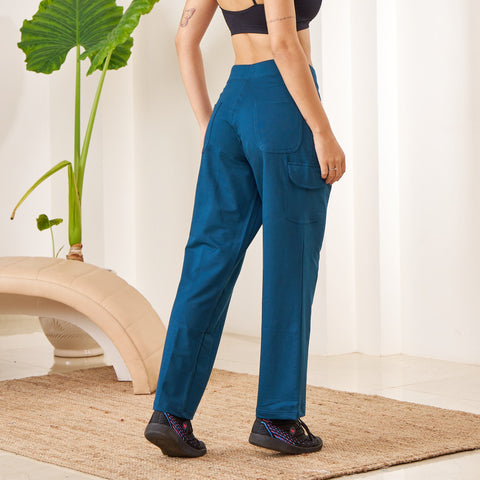 Woman wearing a black sports bra and blue Essential Cargo Pants standing in a minimalistic room with a plant.