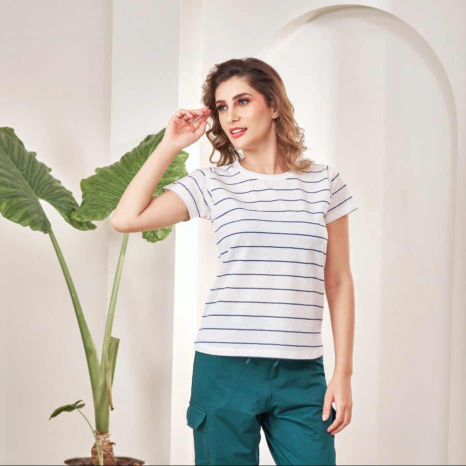 Woman wearing a striped shirt and green pants standing next to a plant in a minimalistic setting