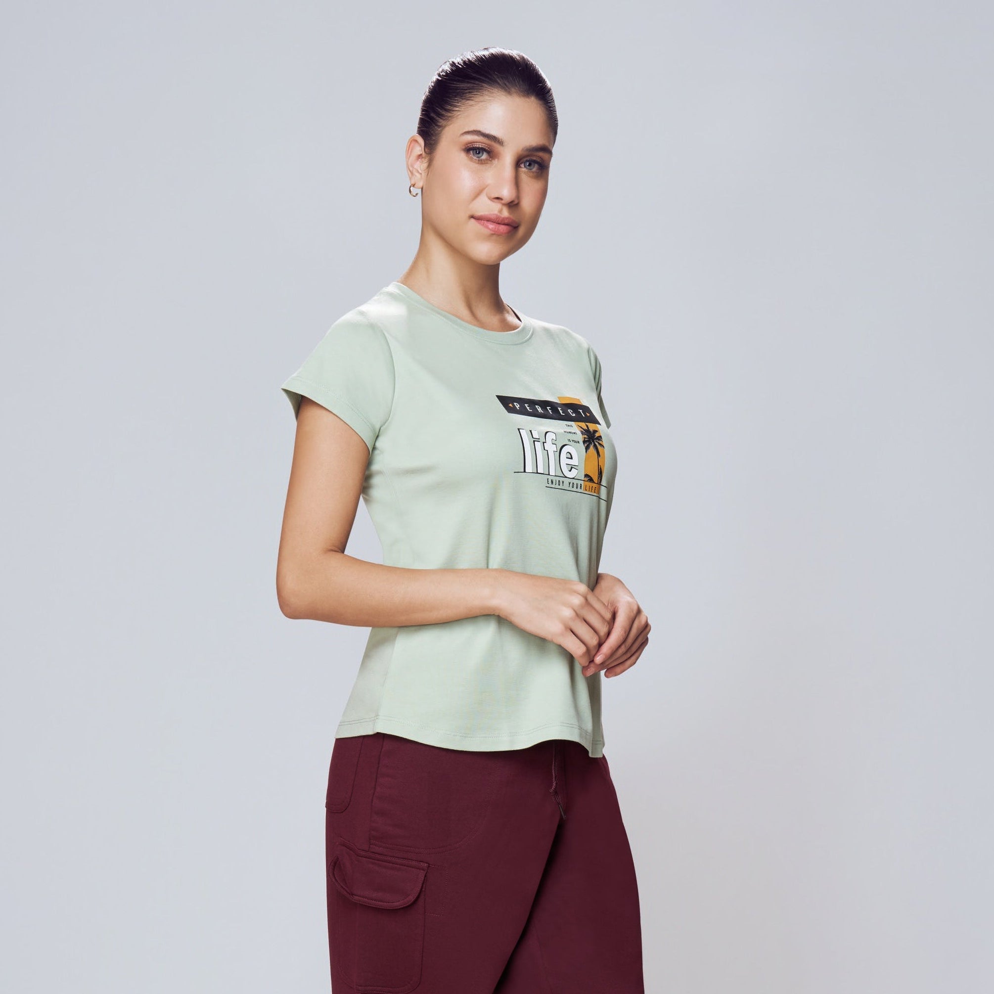 Woman wearing a light green t-shirt with a logo and maroon pants on a white background