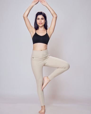 Woman in black sports bra and beige leggings performing a yoga pose against a plain background