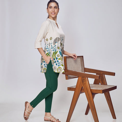 Woman in a floral top and green pants standing next to a wooden chair on a white background