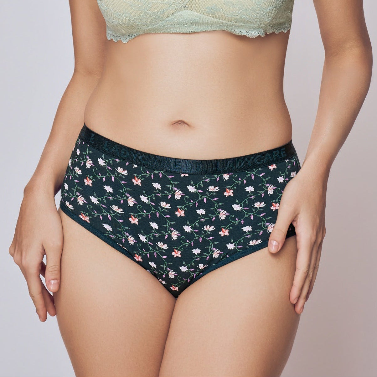 Woman wearing a light green bandeau top and dark patterned high-waisted bikini bottoms on a plain background