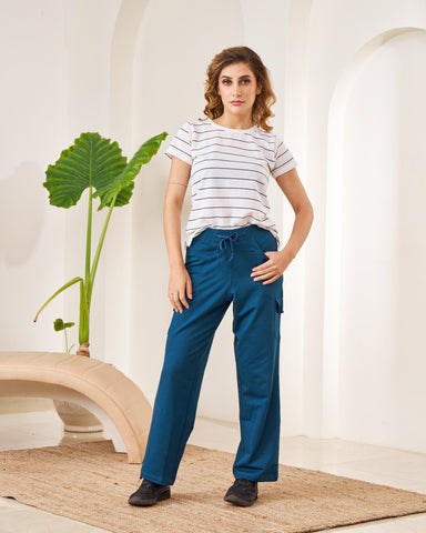 Woman wearing a striped shirt and blue pants standing in a minimalistic room with white walls and a plant.