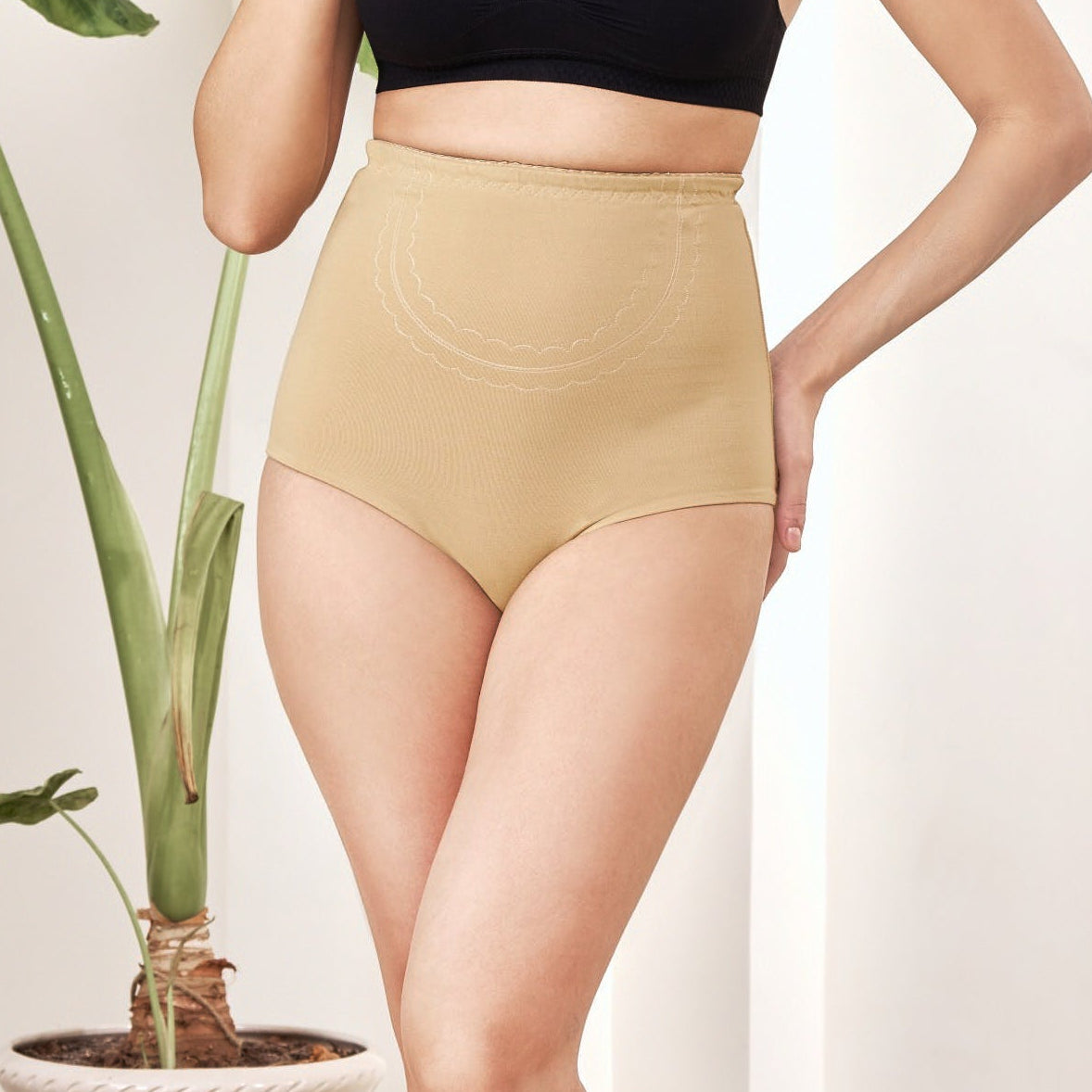 Woman in black sports bra and beige high-waisted shorts standing in a modern interior setting with a plant.