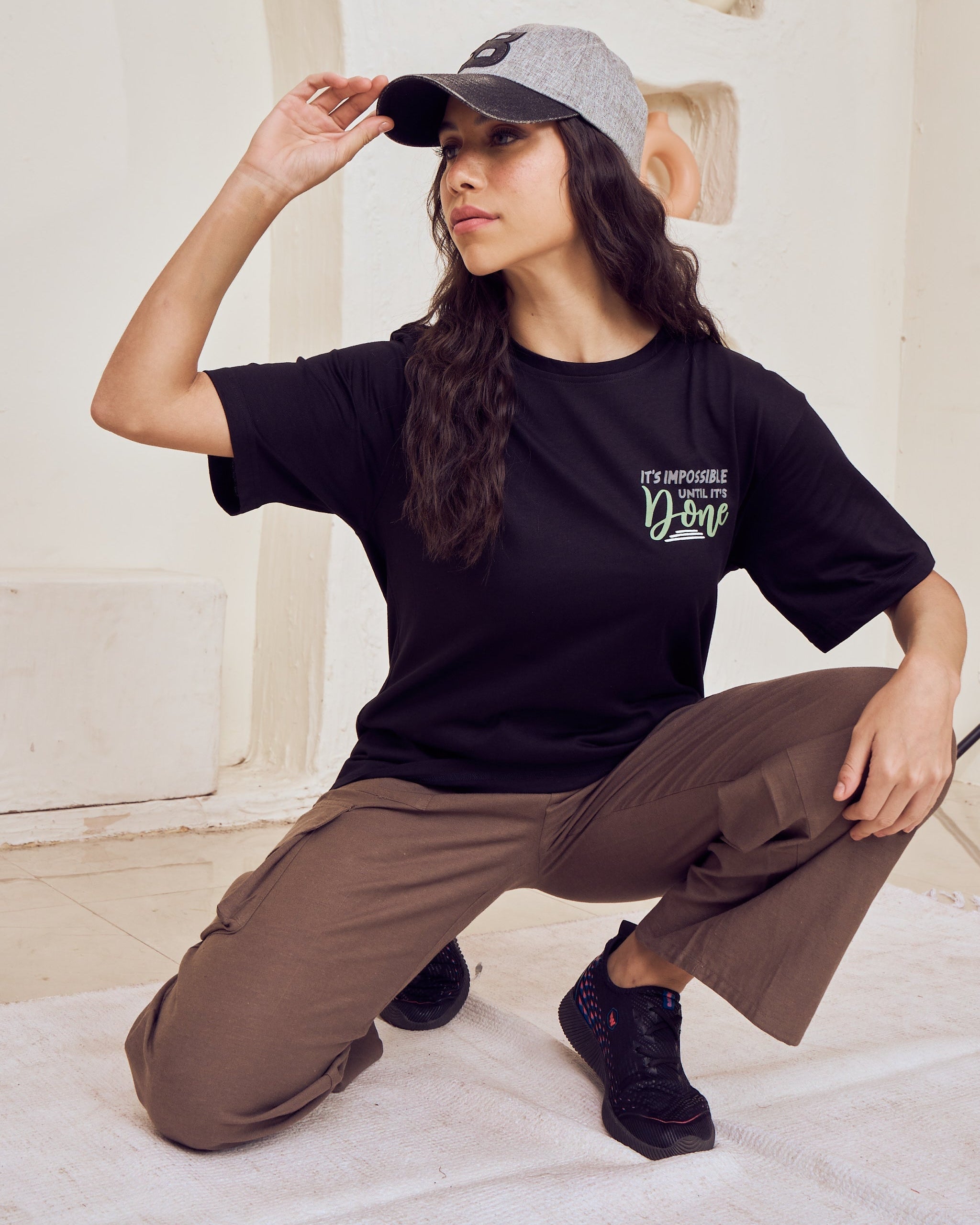 Woman wearing a black t-shirt and brown pants, posing against a textured wall.