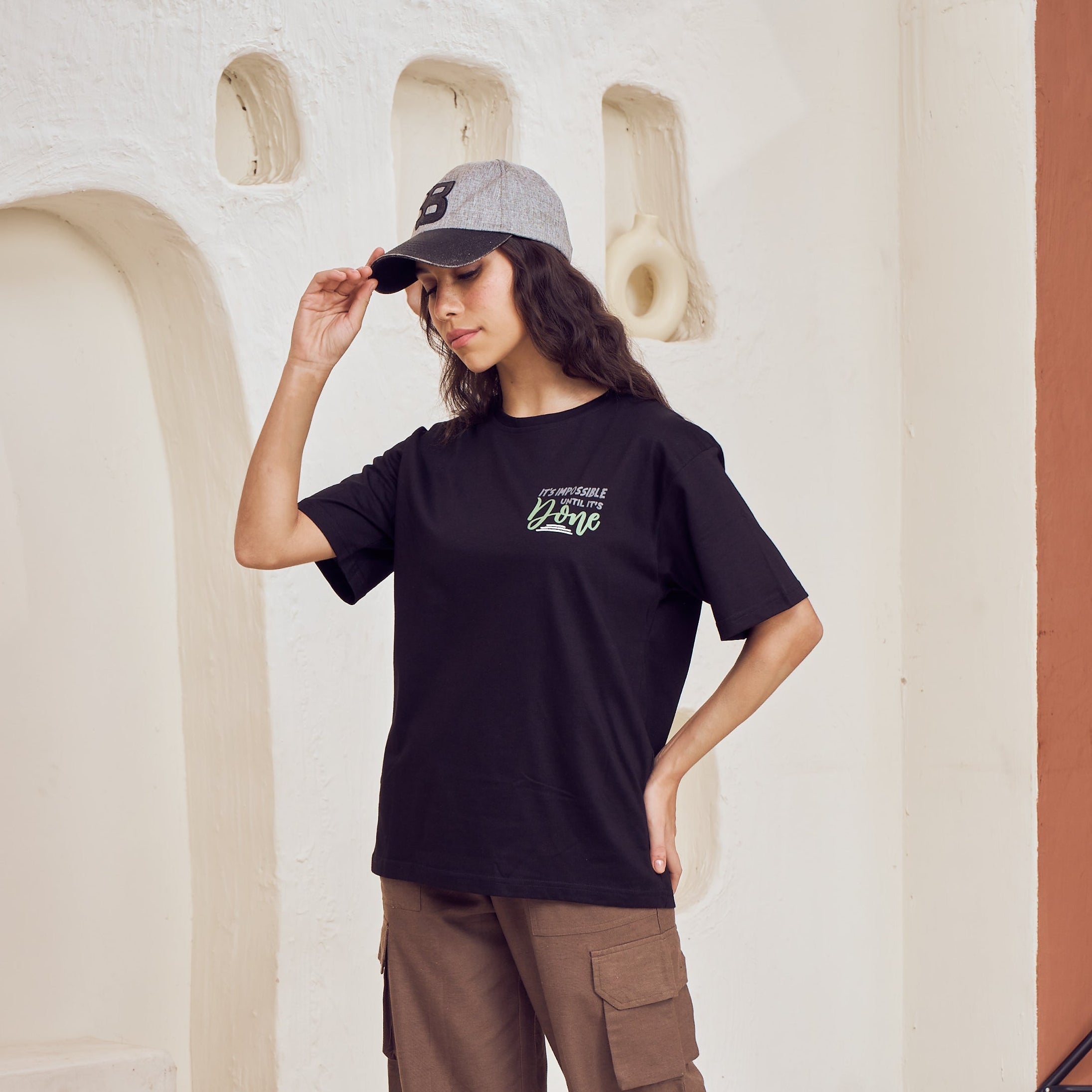 Woman wearing a black t-shirt with a logo and brown cargo pants, standing in a minimalistic indoor setting.