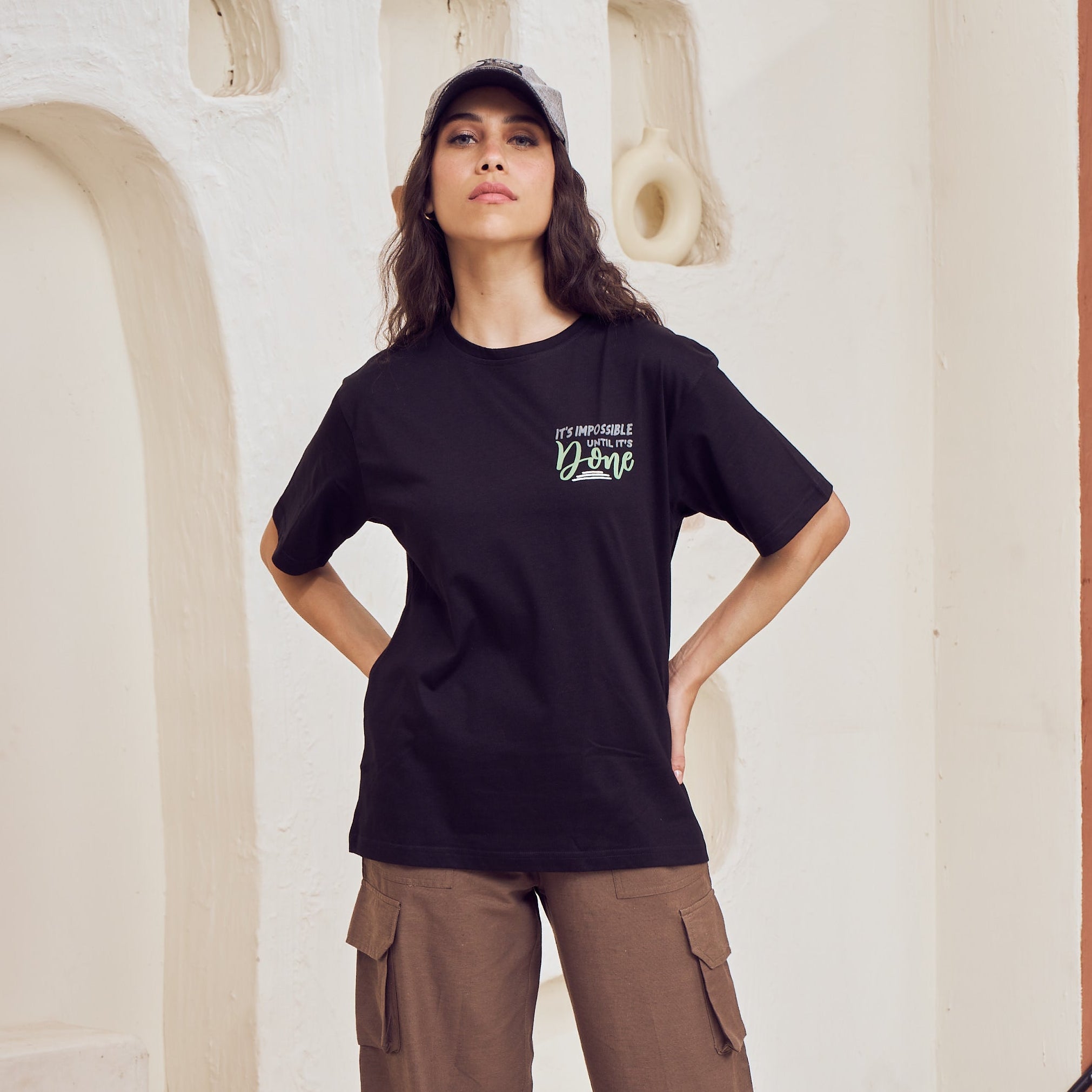 Person wearing a black t-shirt with a logo and brown pants in a modern indoor setting