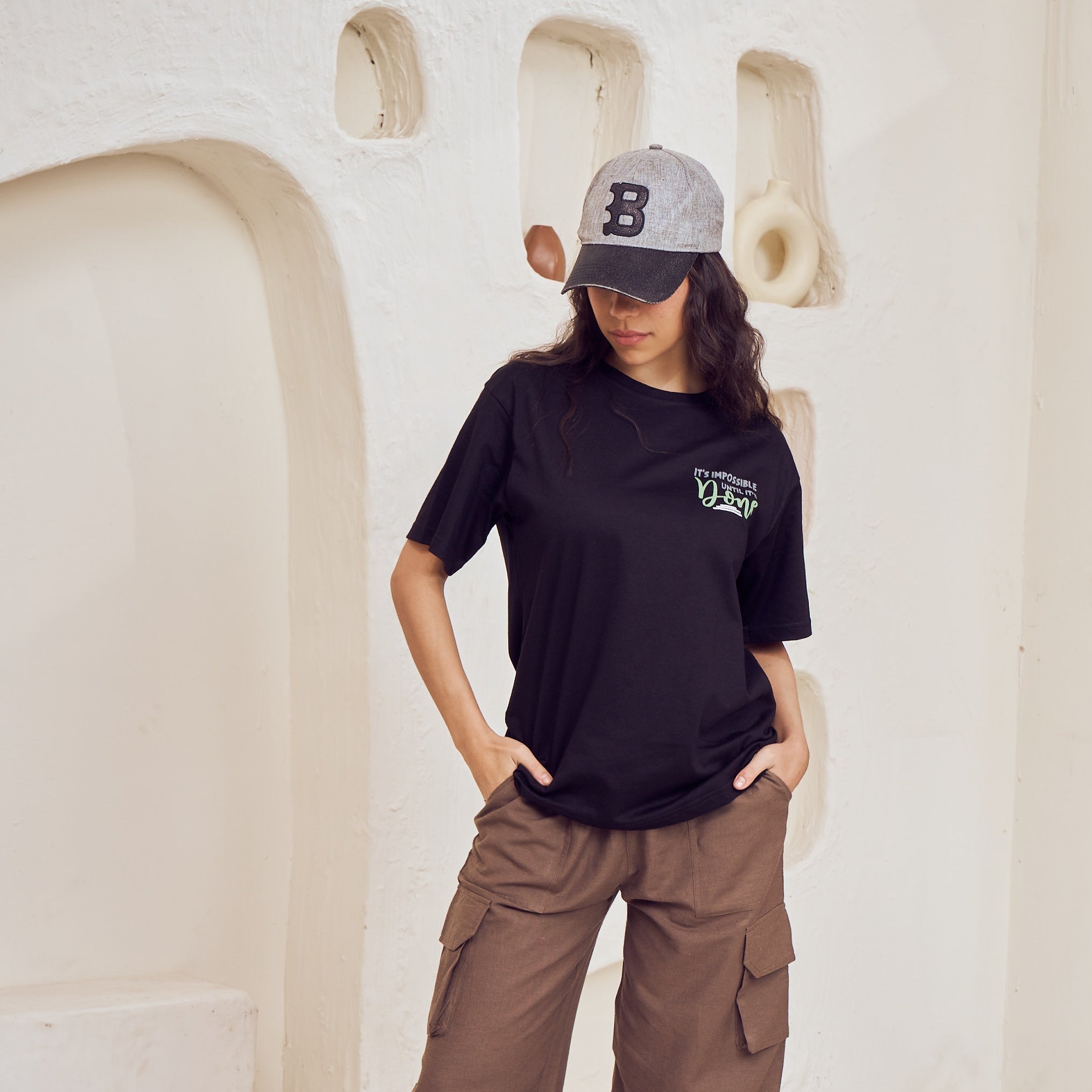 Person wearing a black t-shirt with a logo and brown cargo pants in a minimalistic room.