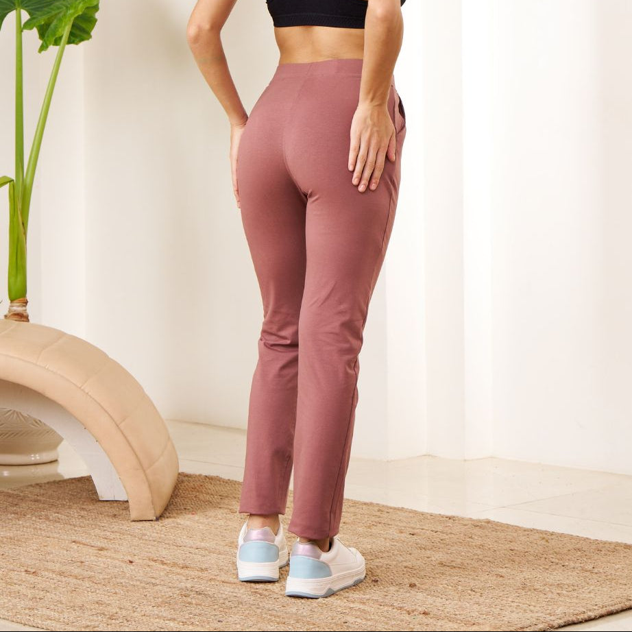 Woman wearing a black sports bra and pink Power Move Track Pants standing in a minimalistic room with a plant.