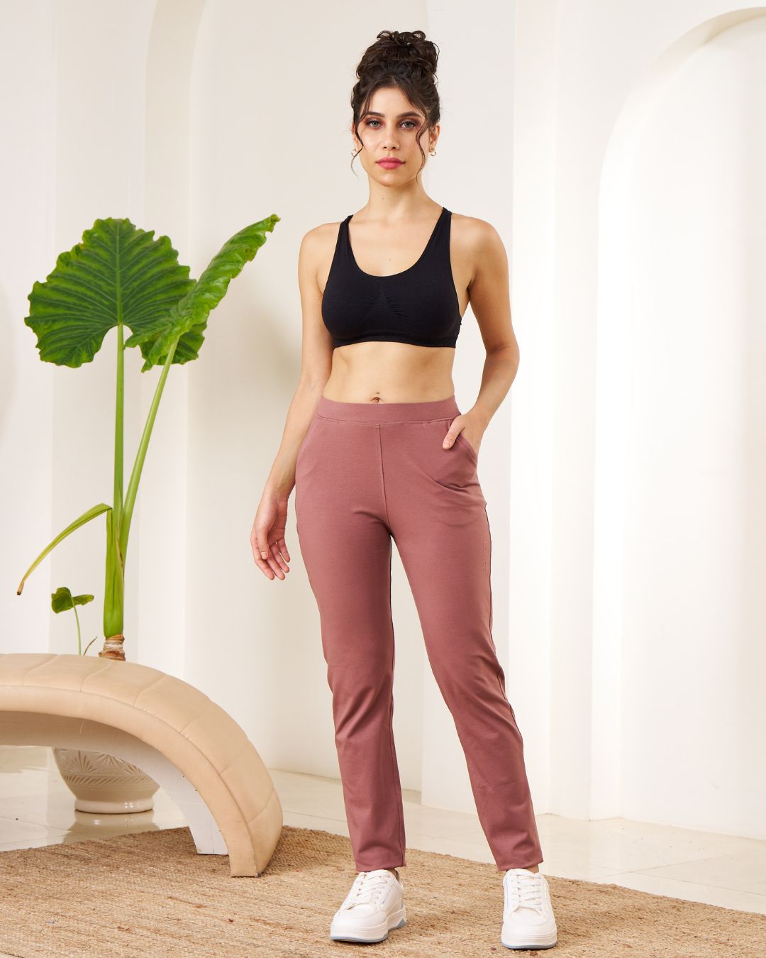 Woman wearing a black sports bra and pink leggings standing indoors with a plant in the background.