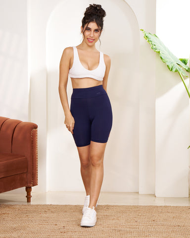Woman in white sports bra and navy bike shorts standing in a room with a brown couch and white wall.