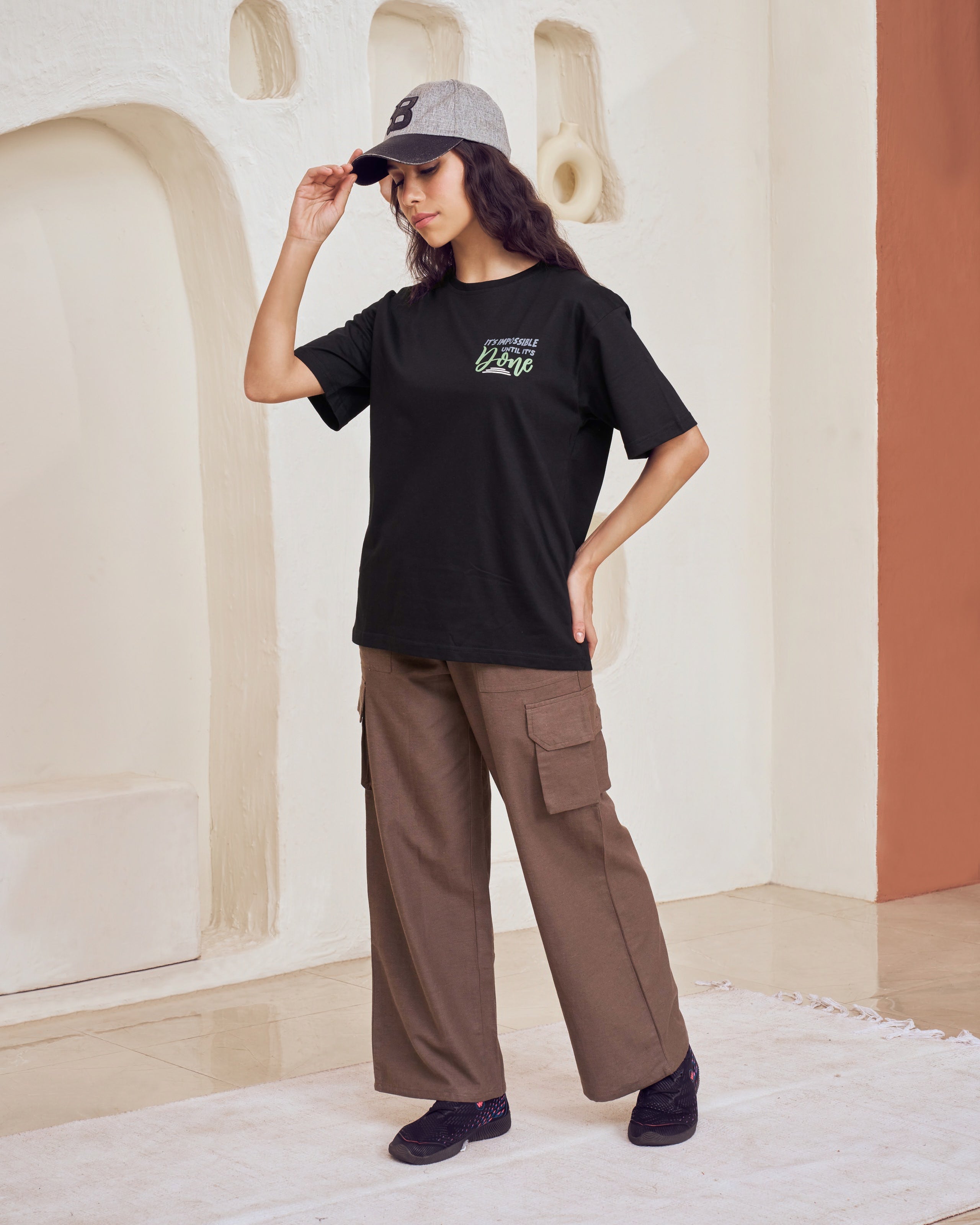 Person wearing a black t-shirt with a logo and brown cargo pants in a modern interior setting.